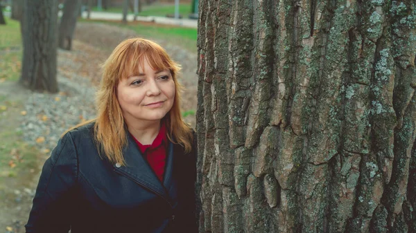 Una mujer seria mirando desde detrás del árbol. Mujer bastante tranquila tocando el árbol y mirando a la cámara en el bosque tranquilo — Foto de Stock