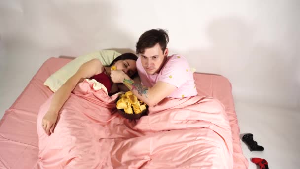 Młoda para leży w łóżku, ogląda film i je chipsy. Mąż karmi żonę żetonami. Kobieta zasypia z nudnego filmu.. — Wideo stockowe