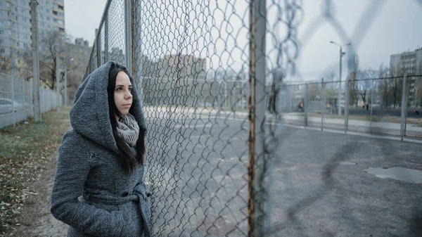 Uma jovem mulher bonita no casaco cinza posa perto de uma cerca de treliça em um final de outono . — Fotografia de Stock