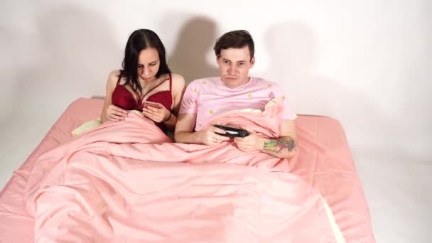 Ein junges Ehepaar in einem Bett. Ein gutaussehender Mann spielt ein Videospiel. Eine schöne Frau blättert mit einem Handy durch die verschiedenen Nachrichten im sozialen Netzwerk. — Stockvideo