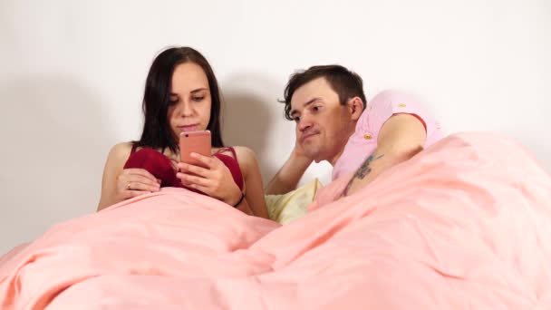 Um jovem casal está deitado em uma cama. A esposa está lançando através das várias notícias em um telefone móvel. O marido está olhando para sua esposa com descontentamento, porque ela não prestar atenção a ele. — Vídeo de Stock