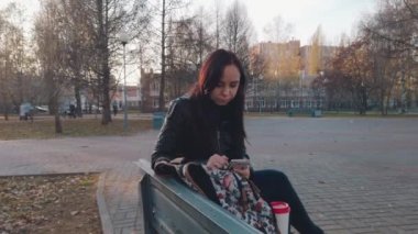 Sırt çantalı genç ve güzel bir kadın bankta oturuyor ve parkta kahve içiyor..