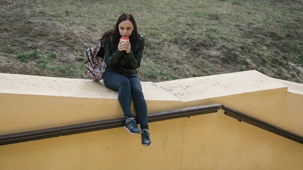 Μια νεαρή όμορφη γυναίκα με καθημερινά ρούχα κάθεται σε ένα τσιμεντένιο φράχτη, κοιτάζει αλλού και κρατάει ένα φλιτζάνι καφέ.. — Φωτογραφία Αρχείου