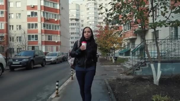 En ung vacker kvinna i casual kläder med en ryggsäck på axeln går och dricker en kaffe på gården i staden. — Stockvideo