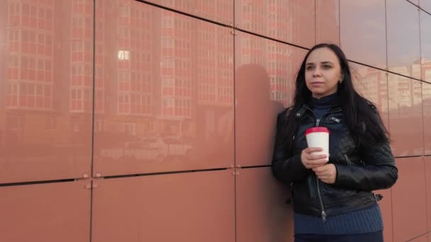 Una joven mujer guapa con ropa casual se encuentra cerca de la pared de azulejos y bebiendo un café.. — Vídeos de Stock