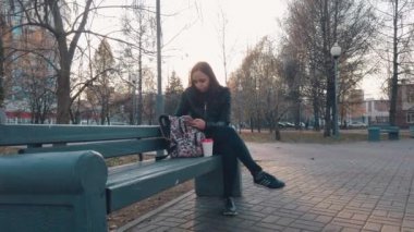Sırt çantalı genç ve güzel bir kadın bankta oturuyor ve parkta kahve içiyor..