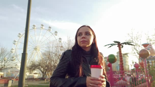 Una joven mujer guapa con ropa casual con mochila está sentada en un banco y bebiendo un café en el parque.. — Vídeos de Stock