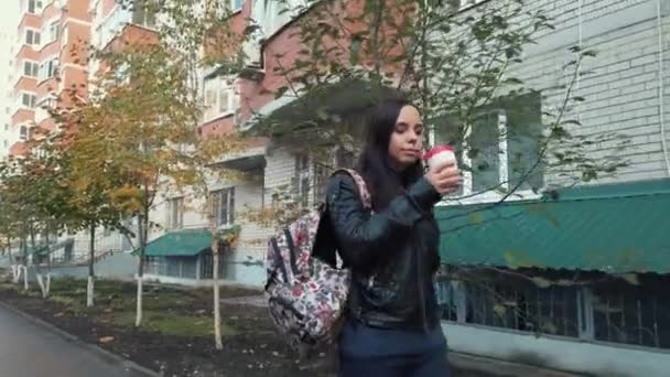 Een jonge mooie vrouw in casual kleding met een rugzak op haar schouder gaat en drinkt een koffie op de binnenplaats van de stad. — Stockvideo