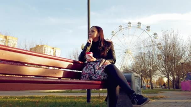 Een jonge mooie vrouw in casual kleding met een rugzak zit op een bankje en drinkt een koffie in het park. — Stockvideo
