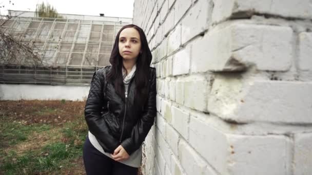 Kvinna i svart skinnjacka. Tråkig kvinna som tittar åt sidan. ung kvinna i svart läder jacka i bakgrunden av en tegelvägg — Stockvideo