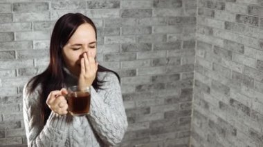 Sıcak kazak giymiş güzel bir kadın ve elinde bir kupa sıcak çayla tuğla duvara doğru esniyor..