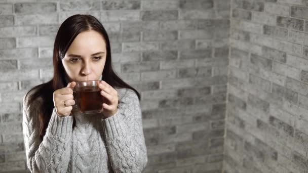 Una joven hermosa mujer en un suéter gris bebe un té, contra la pared de ladrillo gris — Vídeos de Stock