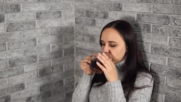 Una joven hermosa mujer en un suéter gris bebe un té, contra la pared de ladrillo gris — Vídeos de Stock