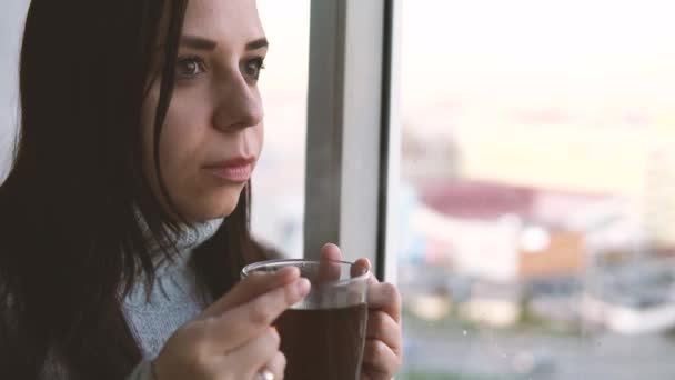 Een jonge mooie vrouw in een grijze trui drinkt een thee, kijkend uit het raam op het balkon. — Stockvideo