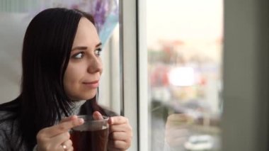 Gri kazaklı güzel bir kadın çay içer, balkondan dışarı bakar..