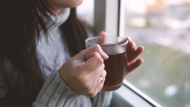 灰色のセーターを着た若い美しい女性がお茶を飲み バルコニーの窓から外を見る — ストック動画
