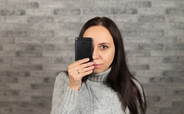 Uma jovem mulher bonita fecha os olhos com um telefone. A mulher está escondida atrás de um telemóvel. O conceito de olhar através do prisma da tela . — Fotografia de Stock