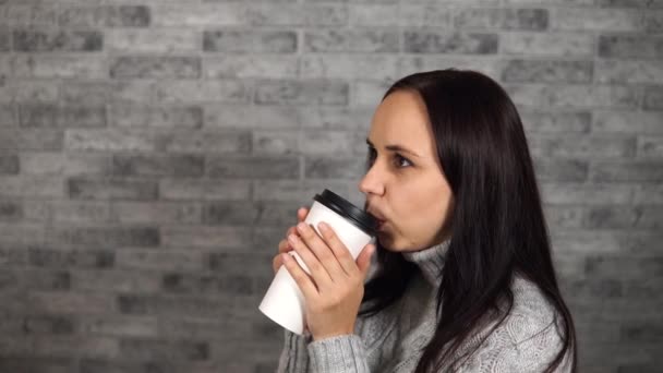 Eine junge schöne Frau im grauen Pullover trinkt einen Kaffee vor dem Hintergrund einer grauen Ziegelwand. — Stockvideo