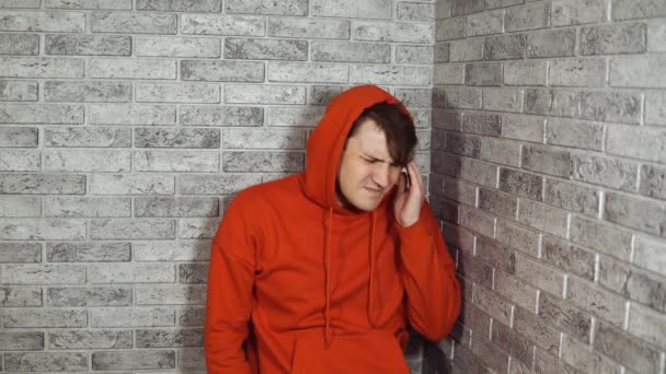 Um homem novo em um hoodie vermelho está prendendo suas mãos em temples na dor em um fundo do tijolo. — Vídeo de Stock