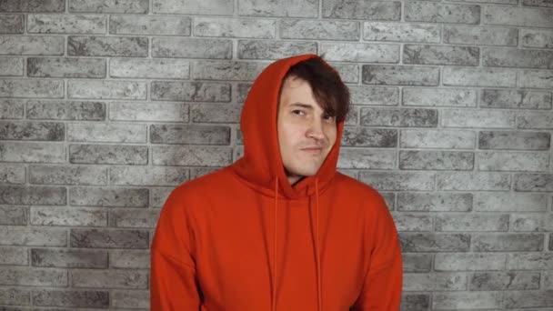 Um homem bonito novo em um hoodie vermelho com winks bristles com boca aberta em um fundo cinzento do tijolo. — Vídeo de Stock