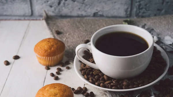 Une tasse de café chaude avec les grains de café et de petits cupcakes sur la table. Composition d'un délicieux café et des collations sucrées . — Photo