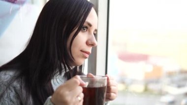 Gri kazaklı güzel bir kadın çay içer, balkondan dışarı bakar..