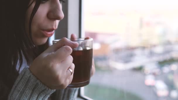 Nahaufnahme, Frau im grauen Pullover trinkt einen Tee und schaut aus dem Fenster auf den Balkon. — Stockvideo