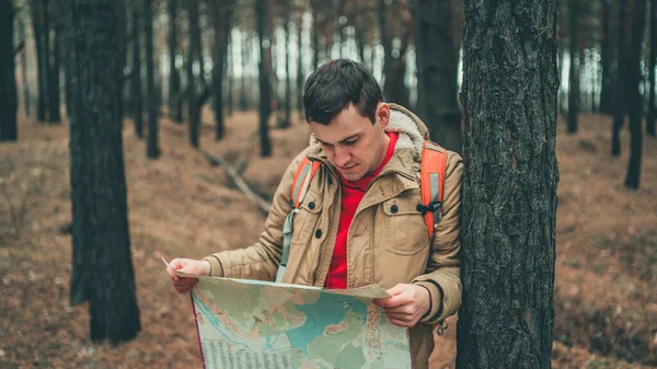 En resande man med karta i skogen. Ett porträtt av den stilige mannen med ryggsäck, stående nära ett träd i kallt väder. — Stockfoto