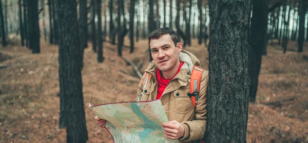 Un hombre viajero con mapa en el bosque. Un retrato del hombre guapo con una mochila, de pie cerca de un árbol en un clima frío . — Foto de Stock