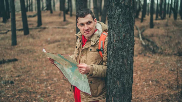 Un hombre viajero con mapa en el bosque. Un retrato del hombre guapo con una mochila, de pie cerca de un árbol en un clima frío . — Foto de Stock
