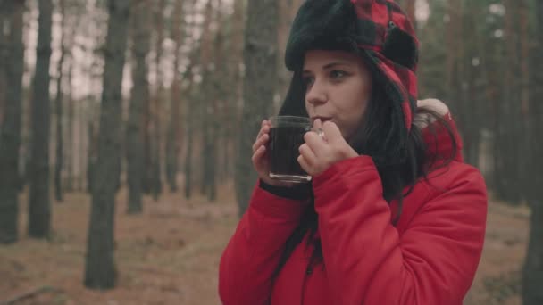 숲 속에서 차를 마시고 있는 아름다운 여인의 초상화. 재킷을 입고 귀덮개를 쓴 모자를 쓴 여자가 추운 날씨에 차를 마시고 있습니다. — 비디오