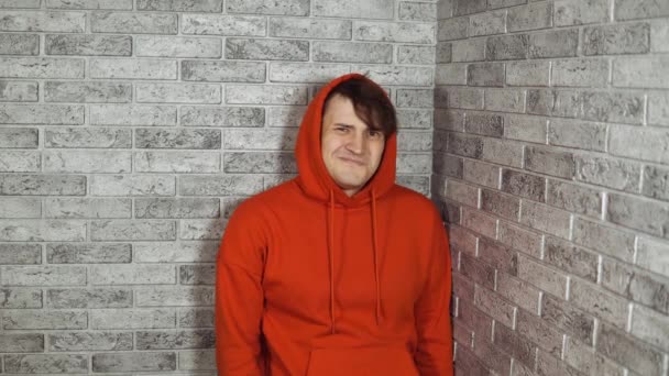 Um homem emocional bonito em um hoodie vermelho mostra polegares para baixo. Um macho mostra um gesto de desaprovação em um fundo de tijolo. O conceito de gostar ou não gostar. — Vídeo de Stock