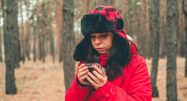 Ormanda bir fincan çayla güzel bir kadının portresi. Ceketli ve kulaklık takan kadın soğuk havada çay içiyor.. — Stok fotoğraf
