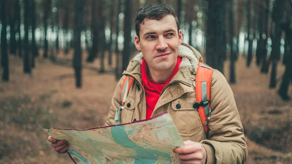 Un hombre viajero con mapa en el bosque. Un retrato del hombre guapo con una mochila, de pie cerca de un árbol en un clima frío . — Foto de Stock