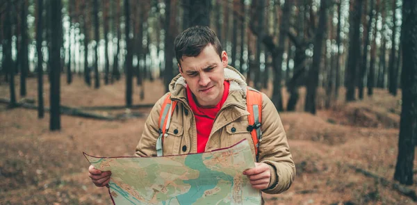 A traveling man with map in woods. A portrait of the handsome man with a backpack, standing near a tree in a cold weather.