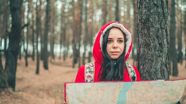 Uma mulher viajante com mapa na floresta. Um retrato da mulher bonita com uma mochila, de pé perto de uma árvore em um tempo frio. — Fotografia de Stock