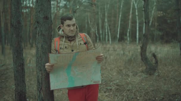 Egy utazó ember térképpel az erdőben. Egy portré a jóképű férfiról hátizsákkal, aki egy fa mellett áll hideg időben.. — Stock videók