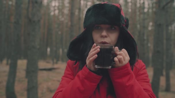 Ein Porträt der hübschen Frau mit einer Tasse Tee im Wald. Die Frau in Jacke und Hut mit Ohrenklappen trinkt bei kaltem Wetter einen Tee. — Stockvideo