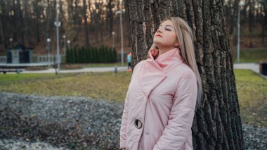 Bir kadın ağacın arkasından dışarı bakıyor. Sakin bir kadın ağaca dokunuyor ve sessiz ormanda kameraya bakıyor.