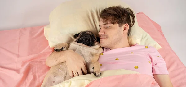 Un bel homme allongé avec un carlin au lit. Jeune homme dans un lit sous un tapis avec son chien dans une étreinte. Beau jeune hipster avec son carlin reposant sur le lit à la maison — Photo