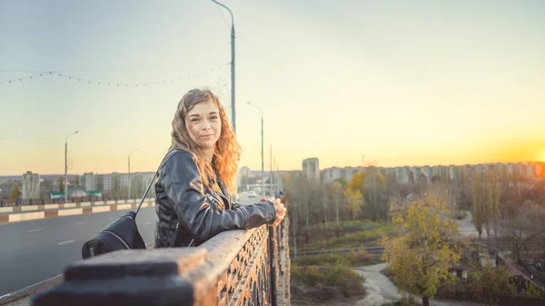 一个身穿皮夹克、背着黑色背包的年轻漂亮的女人站在桥上，面对着市区的背景微笑着. — 图库照片