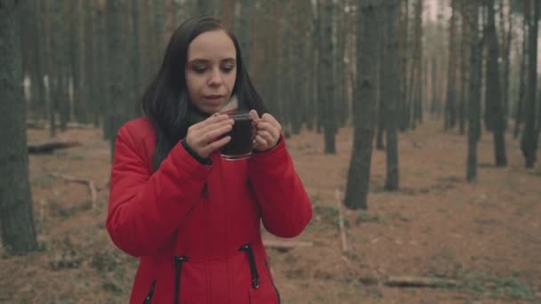 Egy csinos fiatal nő alkalmi ruhában teát iszik, hogy melegen tartsa magát hideg időben az erdőben.. — Stock videók