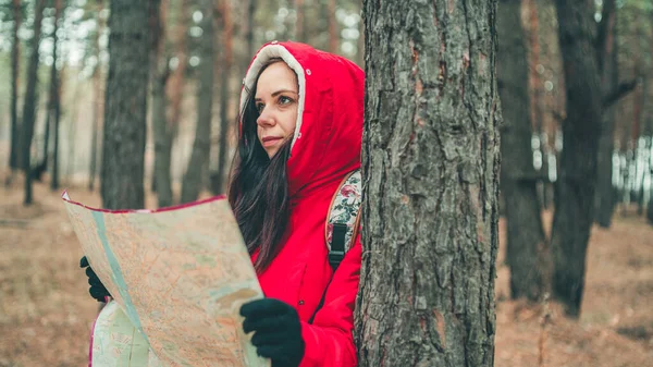 A traveling woman with map in woods. A portrait of the pretty woman with a backpack, standing near a tree in a cold weather.