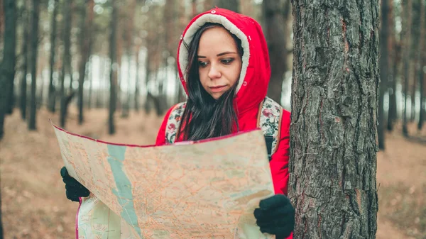 A traveling woman with map in woods. A portrait of the pretty woman with a backpack, standing near a tree in a cold weather. — Stock Photo, Image