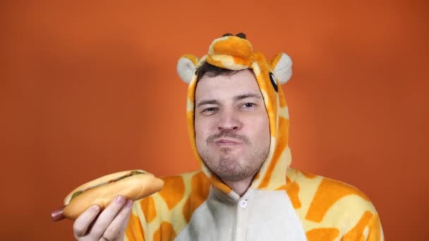 En stilig man i en stor pyjamas av giraff äter en varmkorv på orange bakgrund i studion.. — Stockvideo