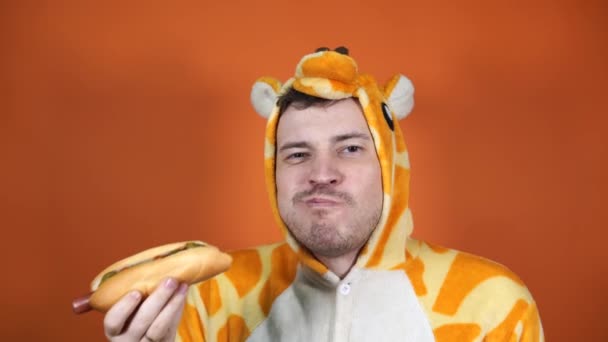 Un bel homme dans un grand pyjama de girafe mange un hot dog sur fond orange en studio. — Video