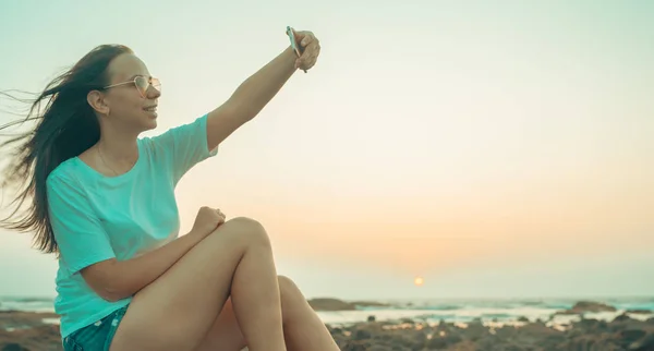 Uma bela jovem está tomando selfie em um telefone celular, sentado nas rochas junto ao mar contra o pôr do sol . — Fotografia de Stock