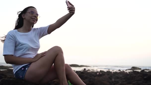 Una hermosa joven se está tomando selfie en un teléfono móvil, sentado en las rocas junto al mar contra el atardecer . — Vídeo de stock