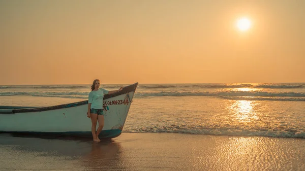 Uma jovem mulher está perto do barco junto ao mar contra o pôr do sol . — Fotografia de Stock