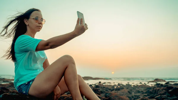 Девушка делает селфи на смартфоне, сидя на пляже на фоне вечернего неба — стоковое фото